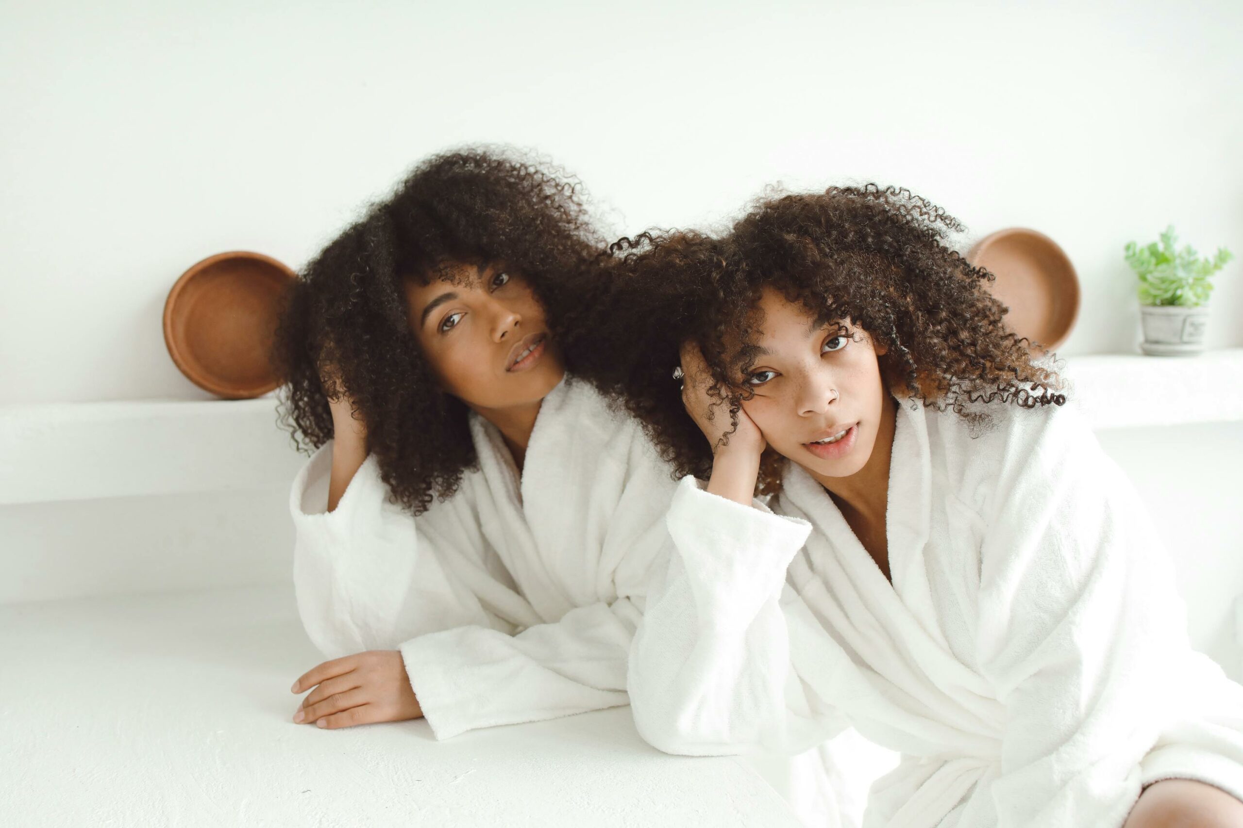 Two African American women in white bathrobes with curly hair, looking relaxed indoors.